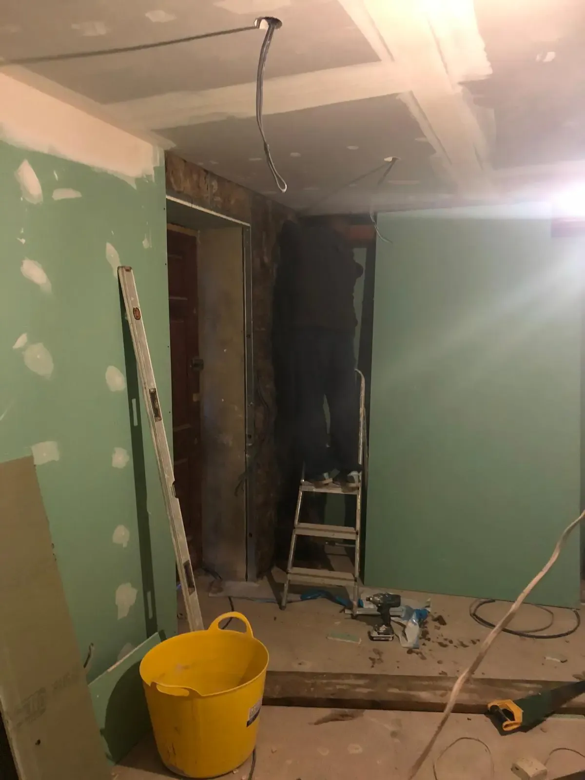 Moisture-resistant plasterboard installed and joint-taped on walls and ceiling, with a decorator finishing the reskim from a step ladder